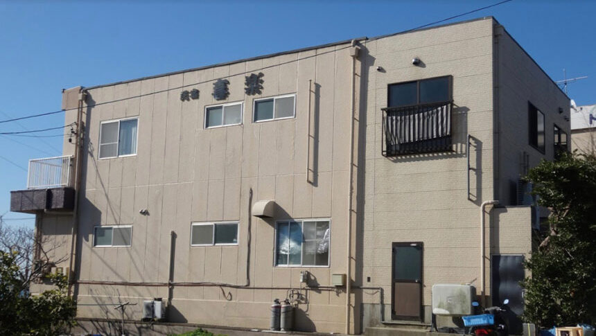 愛知県南知多の民宿 喜楽（きらく）の外観写真。ベージュのサイディング外壁に「民宿 喜楽」と黒い文字で書かれている。2階建ての建物で、左側には小さなバルコニーがあり、1階部分にはプロパンガスのボンベや入り口のドアが見える。快晴の空を背景にした、静かで落ち着いた佇まい