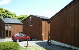 静岡県・南伊豆の犬と泊まれる貸別荘「小さなリゾートハウス」｜緑豊かな森を背に並んで建つ、茶色の木目調がおしゃれな3棟のコテージ。手前の砂利敷きのスペースには赤いコンパクトカーが停まっている。