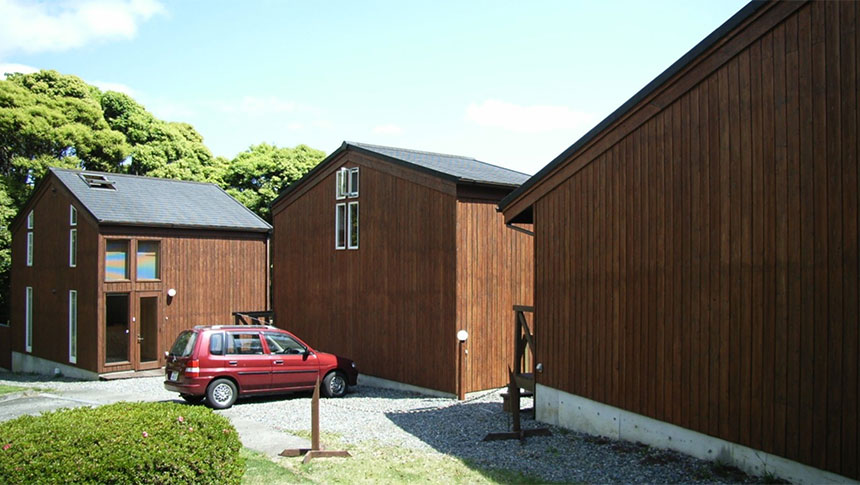 静岡県・南伊豆の犬と泊まれる貸別荘「小さなリゾートハウス」｜緑豊かな森を背に並んで建つ、茶色の木目調がおしゃれな3棟のコテージ。手前の砂利敷きのスペースには赤いコンパクトカーが停まっている。