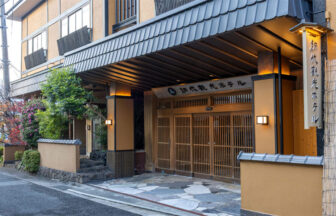 網代観光ホテルの正面玄関。温かみのあるオレンジ色の壁に、重厚な黒い瓦屋根が特徴的。入り口は格子状の木製引き戸で、床面には石畳が敷かれている。右側には「網代観光ホテル」と縦書きされた木製看板がライトアップされている｜静岡県熱海の犬と泊まれる宿｜網代観光ホテル