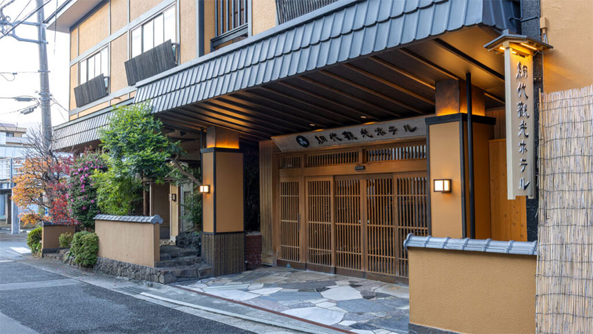 網代観光ホテルの正面玄関。温かみのあるオレンジ色の壁に、重厚な黒い瓦屋根が特徴的。入り口は格子状の木製引き戸で、床面には石畳が敷かれている。右側には「網代観光ホテル」と縦書きされた木製看板がライトアップされている｜静岡県熱海の犬と泊まれる宿｜網代観光ホテル