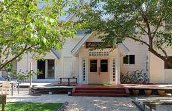 静岡県・下田の犬と泊まれる宿「アーネストハウス」｜青々とした木の葉越しに見える、洋風な建物の正面玄関。白い外壁に薄いピンク色の両開きドアがあり、その上には宿の看板が掲げられています。玄関の右側には数台の自転車が停まっており、左側には屋外テーブルと椅子が置かれたテラススペースがあります。地面は石畳で、木漏れ日が美しく模様を描いています。