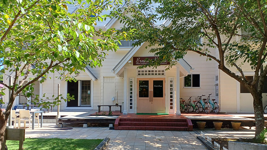 静岡県・下田の犬と泊まれる宿「アーネストハウス」｜青々とした木の葉越しに見える、洋風な建物の正面玄関。白い外壁に薄いピンク色の両開きドアがあり、その上には宿の看板が掲げられています。玄関の右側には数台の自転車が停まっており、左側には屋外テーブルと椅子が置かれたテラススペースがあります。地面は石畳で、木漏れ日が美しく模様を描いています。