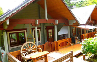 静岡県・伊豆高原の犬と泊まれる宿「高原のリゾートログハウス凛灯」｜大きなウッドデッキがある平屋のログハウス。薄いグレーの壁に赤茶色の梁が映え、デッキには車輪を背もたれにした木製のベンチが置かれています。屋根は急勾配で、軒下には繊細な装飾が施されています