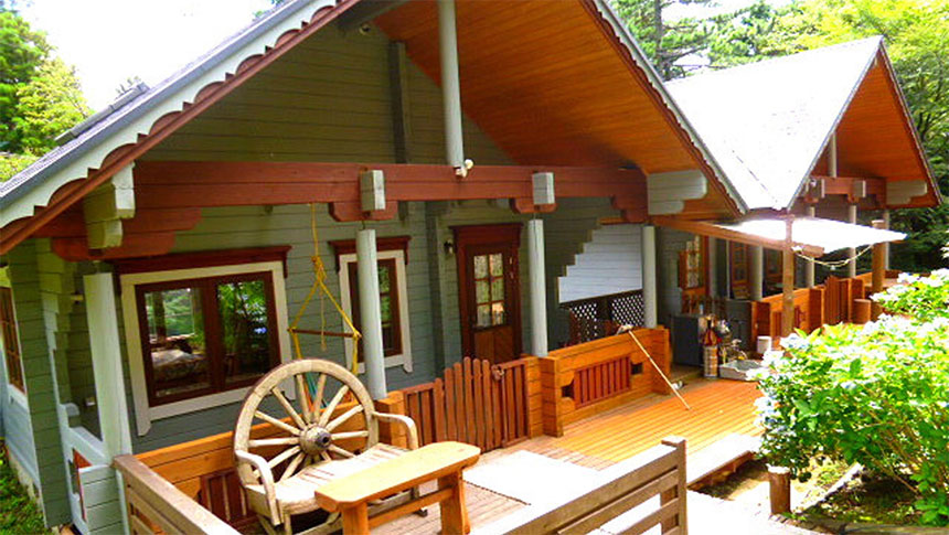 静岡県・伊豆高原の犬と泊まれる宿「高原のリゾートログハウス凛灯」｜大きなウッドデッキがある平屋のログハウス。薄いグレーの壁に赤茶色の梁が映え、デッキには車輪を背もたれにした木製のベンチが置かれています。屋根は急勾配で、軒下には繊細な装飾が施されています