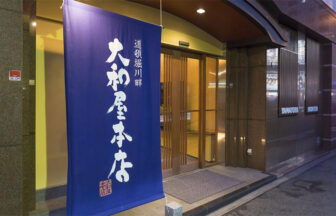 大阪・道頓堀川畔にある犬と泊まれる宿「大和屋本店」の正面玄関。鮮やかな紺色の大きな日除け暖簾（のれん）に白い文字で「大和屋本店」と記された、伝統ある和風旅館の入り口の様子