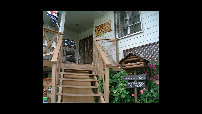 兵庫県淡路市にある犬と泊まれる宿「ペンションZOO」のエントランス。木製の階段の先にアットホームな玄関があり、看板には「PENSION ZOO」の文字。階段横には花々が咲き、ログハウス風のポストが置かれた温かみのある外観
