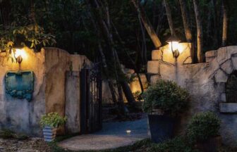 滋賀県大津市北比良にある「オーベルジュ メソン」の夜のエントランス。暖かなアンティークライトに照らされた石造りの壁と、緑色のレリーフ看板、鉄製の門が静かな森の中に佇む様子。1日4組限定の隠れ家宿。