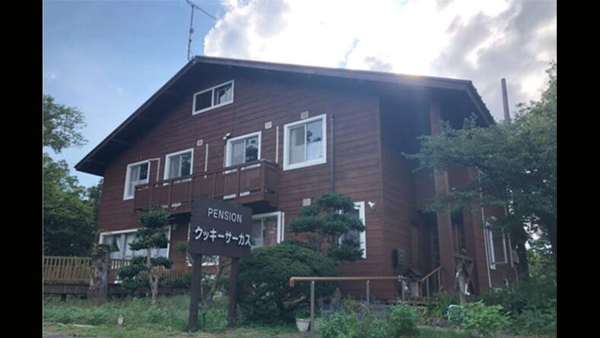 鳥取県・大山のペンション　クッキーサーカス｜緑豊かな木々に囲まれた、落ち着いた茶色の木造2階建てペンション。入り口に立つ手作り感のある看板が目印