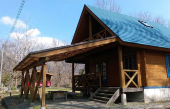 岡山県・蒜山高原の犬と泊まれる「貸別荘ちょっぷ村」｜晴天下、森に囲まれた場所に建つログハウス風の木造コテージ。青い屋根と天窓、ウッドデッキ、隣接する木製の屋根付き駐車スペース（カーポート）があり、奥には別の小さな赤い小屋が見える