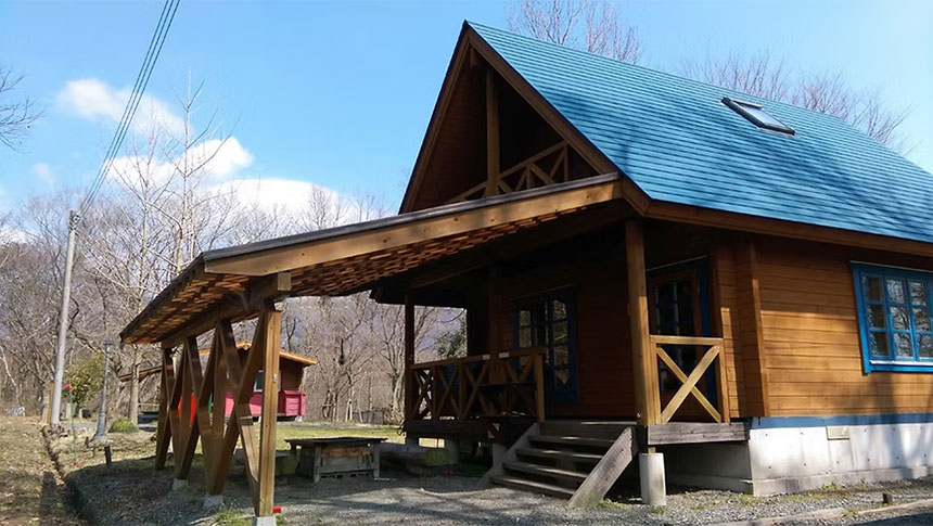 岡山県・蒜山高原の犬と泊まれる「貸別荘ちょっぷ村」｜晴天下、森に囲まれた場所に建つログハウス風の木造コテージ。青い屋根と天窓、ウッドデッキ、隣接する木製の屋根付き駐車スペース（カーポート）があり、奥には別の小さな赤い小屋が見える