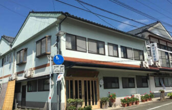 熊本県阿蘇の内牧温泉にある犬と泊まれる宿「村田家旅館」の外観。青空の下、親しみやすい雰囲気の和風旅館で、玄関先には四季折々の花が咲き、看板に「村田家旅館」の文字