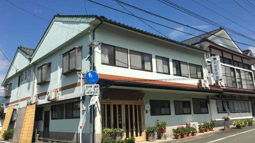 熊本県阿蘇の内牧温泉にある犬と泊まれる宿「村田家旅館」の外観。青空の下、親しみやすい雰囲気の和風旅館で、玄関先には四季折々の花が咲き、看板に「村田家旅館」の文字