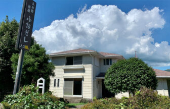 阿蘇の豊かな自然の中に立つ、2階建ての一棟貸し貸別荘「ふなざし」の外観写真。建物は白い横羽目板で、庭の緑豊かな生垣と大きな植栽が前面にある。左側のポールには、黒地に白文字で「犬と泊まる 貸別荘 ふなざし」と書かれた縦長の看板が設置されている。背景には広大な青空に浮かぶダイナミックな積乱雲が広がり、のどかで広々とした阿蘇の環境を感じさせる