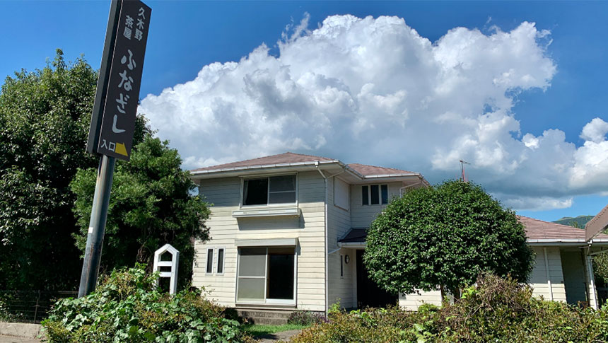 阿蘇の豊かな自然の中に立つ、2階建ての一棟貸し貸別荘「ふなざし」の外観写真。建物は白い横羽目板で、庭の緑豊かな生垣と大きな植栽が前面にある。左側のポールには、黒地に白文字で「犬と泊まる 貸別荘 ふなざし」と書かれた縦長の看板が設置されている。背景には広大な青空に浮かぶダイナミックな積乱雲が広がり、のどかで広々とした阿蘇の環境を感じさせる