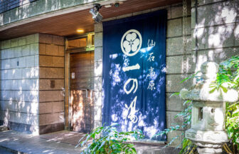 箱根・仙石原にある「仙石高原 一の湯」のエントランス。石造りの外壁に、紺地に白文字で「仙石高原 一の湯」と書かれた大きな暖簾（のれん）が掛かっており、脇には石灯籠と緑の植物が並ぶ趣ある入り口