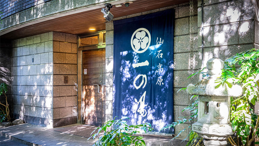 箱根・仙石原にある「仙石高原 一の湯」のエントランス。石造りの外壁に、紺地に白文字で「仙石高原 一の湯」と書かれた大きな暖簾（のれん）が掛かっており、脇には石灯籠と緑の植物が並ぶ趣ある入り口