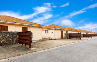 石垣島の名蔵ヴィレッジの外観。快晴の青空を背景に、赤瓦風のオレンジ色の屋根を持つ平屋建ての客室が横一列に並んでいる。建物の前面には沖縄らしい石積みの壁と、プライバシーを守るための木製フェンスが設置されており、南国リゾートの平穏な雰囲気が漂っている｜沖縄県石垣島の犬と泊まれる宿