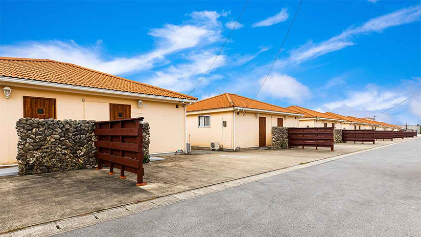 石垣島の名蔵ヴィレッジの外観。快晴の青空を背景に、赤瓦風のオレンジ色の屋根を持つ平屋建ての客室が横一列に並んでいる。建物の前面には沖縄らしい石積みの壁と、プライバシーを守るための木製フェンスが設置されており、南国リゾートの平穏な雰囲気が漂っている｜沖縄県石垣島の犬と泊まれる宿