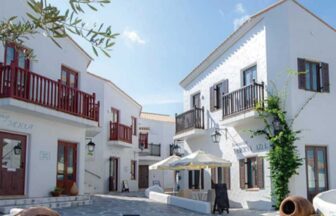 志摩地中海村の白い建物とオレンジ色の瓦屋根が並ぶ石畳の広場。中央には小さな噴水があり、背景に青い空と緑の木々が広がる風景。