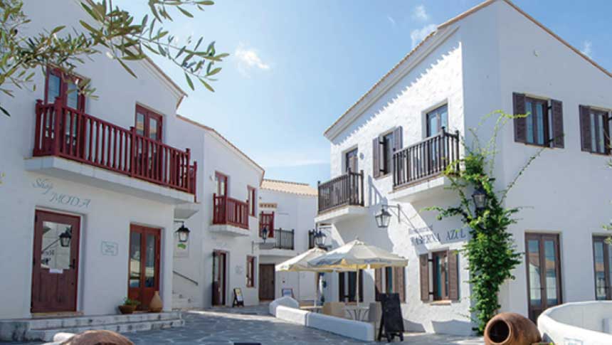 志摩地中海村の白い建物とオレンジ色の瓦屋根が並ぶ石畳の広場。中央には小さな噴水があり、背景に青い空と緑の木々が広がる風景。