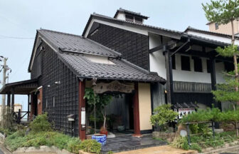 海沿いに建つ、伝統的な蔵造りを思わせる落ち着いた佇まいの旅館「本陣粋月」の外観。曇り空の下、松の木や緑に囲まれた趣のある玄関口が広がる