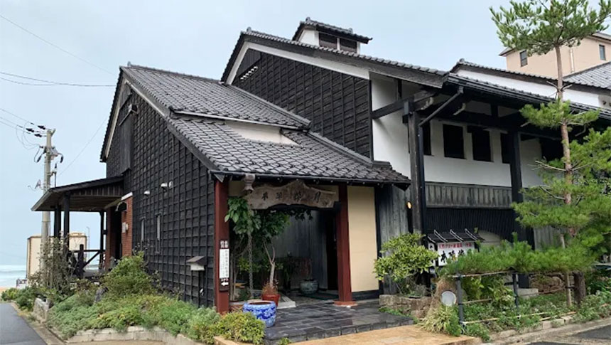 海沿いに建つ、伝統的な蔵造りを思わせる落ち着いた佇まいの旅館「本陣粋月」の外観。曇り空の下、松の木や緑に囲まれた趣のある玄関口が広がる