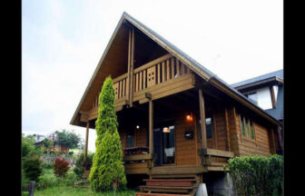 大分県・九重の犬と泊まれる貸別荘「貸別荘飯田T」｜豊かな緑に囲まれた、木のぬくもりを感じる2階建てのログハウスの外観。広いベランダとウッドデッキの階段が特徴的