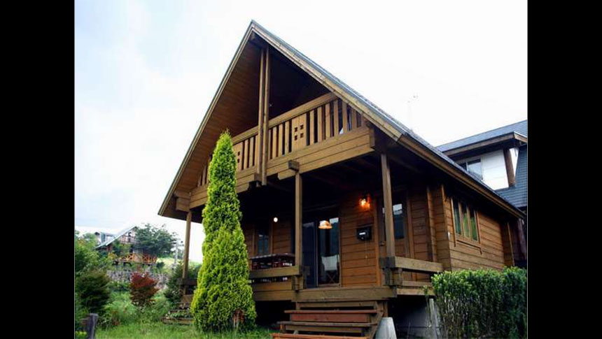 大分県・九重の犬と泊まれる貸別荘「貸別荘飯田T」｜豊かな緑に囲まれた、木のぬくもりを感じる2階建てのログハウスの外観。広いベランダとウッドデッキの階段が特徴的