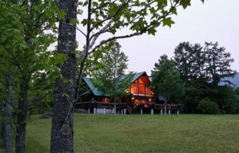 岡山県・蒜山のNadja ゲストハウス｜森と草原に囲まれた静かなログハウス。夕闇の中で室内から漏れる温かな光が印象的なコテージの風景