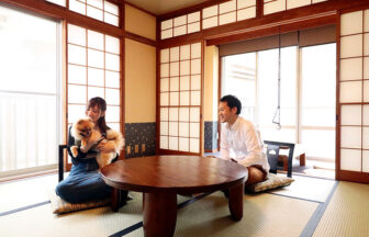 静岡県西伊豆の犬と泊まれる「浜道楽（はまどうらく）」｜伝統的な畳の部屋（和室）にて、丸いちゃぶ台を囲んで座る男女と、女性に抱かれているポメラニアンの様子。部屋は障子窓から明るい光が差し込んでいる