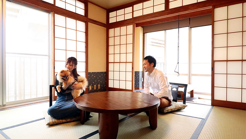 静岡県西伊豆の犬と泊まれる「浜道楽（はまどうらく）」｜伝統的な畳の部屋（和室）にて、丸いちゃぶ台を囲んで座る男女と、女性に抱かれているポメラニアンの様子。部屋は障子窓から明るい光が差し込んでいる