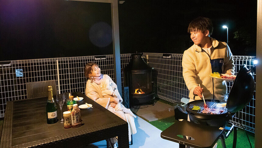 静岡県・伊豆高原の犬と泊まれる宿　ゆがふ「伊豆」｜夜のウッドデッキでBBQを楽しむ男女。男性がグリルでお肉を焼き、女性は椅子に座って愛犬を抱きながら笑顔で見守っている。テーブルにはシャンパンが置かれ、奥には火の灯ったストーブが見える