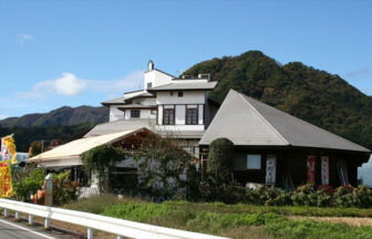 群馬県にある犬と泊まれる宿「ポミエ」の全景。白壁のモダンな建物と、特徴的な大きな三角屋根の木造建築が合わさっており、周囲には緑豊かな山々が広がっている。道路沿いには「りんご狩り」の黄色いのぼり旗がはためいている。