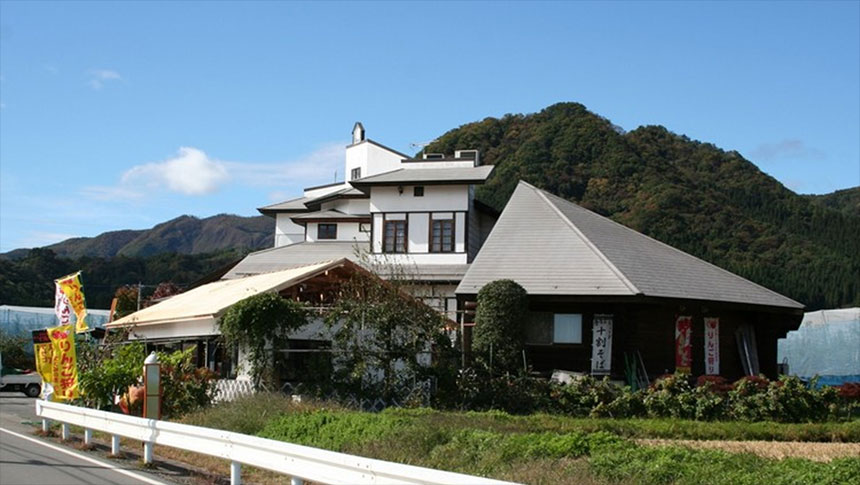 群馬県にある犬と泊まれる宿「ポミエ」の全景。白壁のモダンな建物と、特徴的な大きな三角屋根の木造建築が合わさっており、周囲には緑豊かな山々が広がっている。道路沿いには「りんご狩り」の黄色いのぼり旗がはためいている。