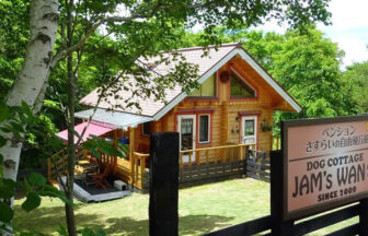 長野県・菅平・峰の原の犬と泊まれる宿「DogコテージJAM’s WAN！」｜夏の緑に囲まれたログハウス風コテージの外観。「JAM'S WAN」と書かれた看板と、専用のドッグランが見える。