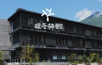 由布院温泉「榎屋旅館」の黒格子の和モダンな外観。青空の下、お宿の背景にそびえる美しい由布岳と、川沿いの静かなロケーション。