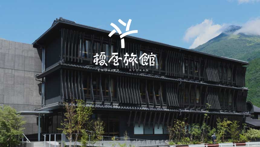 由布院温泉「榎屋旅館」の黒格子の和モダンな外観。青空の下、お宿の背景にそびえる美しい由布岳と、川沿いの静かなロケーション。