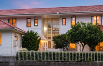 夕暮れ時に温かい明かりが灯る、赤い瓦屋根と白い外壁が特徴的な東伊豆のプチホテル「サザンウインド」の外観。中央の全面ガラスのサンルームと、前庭の白いフェンス、緑の木々が見える。