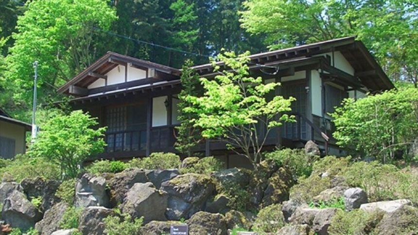 山梨県の愛犬と泊まれる「やすらぎの別邸 四季亭」｜緑豊かな山林を背に、大きな岩が配置された斜面に建つ「やすらぎの別邸 四季亭」の和風コテージの外観。新緑の木々に囲まれた落ち着いた佇まい。