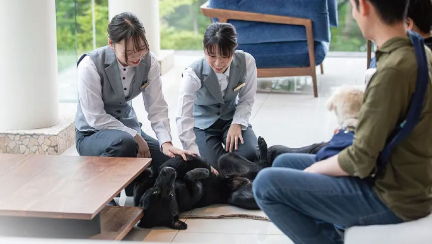 山梨県・山中湖の犬と泊まれる宿「レジーナリゾート山中湖」｜ロビーで笑顔のスタッフに撫でられてリラックスする大きな黒い犬と、それを見守る飼い主。