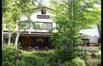群馬県の愛犬と泊まれる宿「ペンションコスモス」｜鮮やかな新緑の木々の合間に佇む、白と茶色を基調としたアットホームな2階建てペンションの外観。右側の木製看板には「Pension コスモス」と書かれており、豊かな自然と調和した温かみのある雰囲気が伝わる。