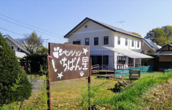 群馬県・北軽井沢の愛犬と泊まれる宿「ペンションいちばん星」｜肉球や星のイラストが描かれた茶色の木製看板があり、奥に白い2階建ての建物とドッグランのような柵が見える、群馬県北軽井沢の愛犬と泊まれる宿の外観