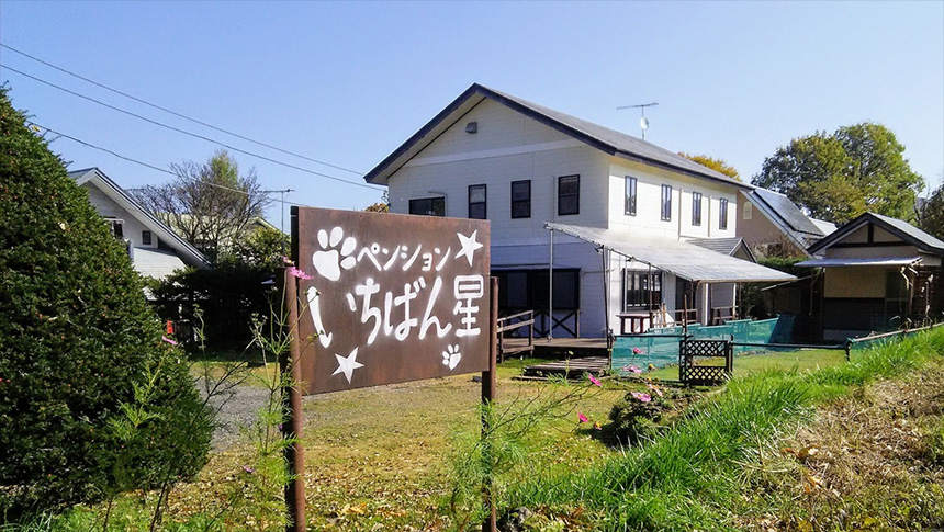 群馬県・北軽井沢の愛犬と泊まれる宿「ペンションいちばん星」｜肉球や星のイラストが描かれた茶色の木製看板があり、奥に白い2階建ての建物とドッグランのような柵が見える、群馬県北軽井沢の愛犬と泊まれる宿の外観