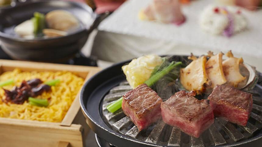 「わんわんパラダイス奈良生駒」の夕食会席より、熱々の鉄板でジュージューと焼かれている厚切りの和牛ステーキ3切れと、殻付きのアワビ、野菜。奥には、お造りの盛り合わせや小鍋、蒸し料理（おこわ）が並んでいる。