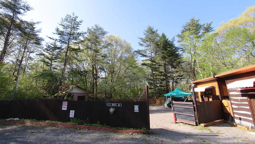 群馬県・北軽井沢の犬と泊まれる宿「ミリーズラブ」｜豊かな緑に囲まれた入り口。ウッドフェンスには「U.S. ROUTE 66」の看板が飾られ、奥には青いタープが設置されたキャンプサイトが見える。