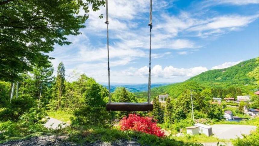 山形県・蔵王温泉にある犬と泊まれる宿「善七の湯」｜高台から蔵王の山並みや街並みを一望できる場所に設置された木製ブランコ。青い空と白い雲、鮮やかな新緑が広がる爽快な風景