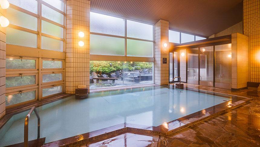 広々とした屋内大浴場。大きなガラス窓からは外の景色が見え、自然光が降り注ぐ中でゆったりと湯船に浸かることができる。