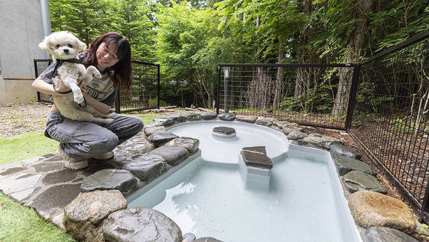 山梨県・山中湖の犬と泊まれる宿「レジーナリゾート山中湖」｜屋外にある石造りの愛犬専用スパ。女性に抱っこされた小型犬が、森の景色を眺めながら温浴を楽しめる施設。
