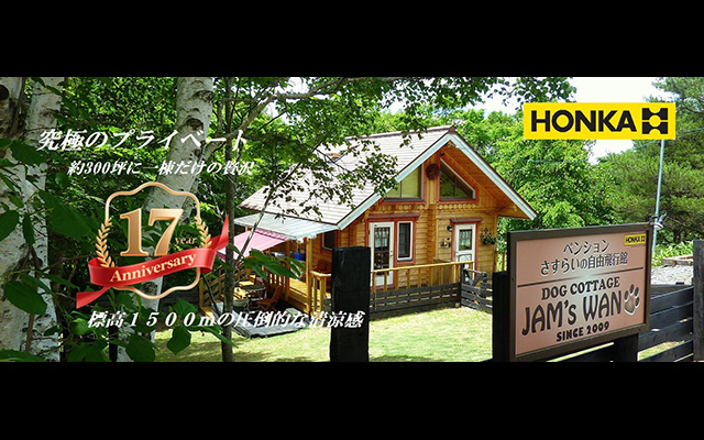 新緑の木々に囲まれた木の温もりを感じる2階建てログハウス。手前には『DOG COTTAGE JAM'S WAN SINCE 2009』と書かれた看板があり、左側には『17 year Anniversary』のエンブレムと『標高1500mの圧倒的な清涼感』というキャッチコピーが添えられている。